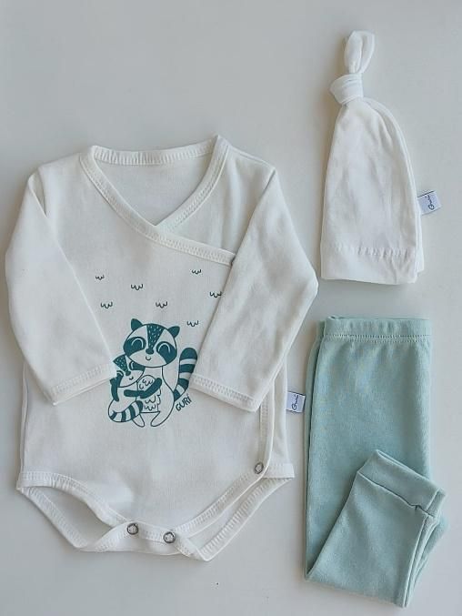 Conjunto de bebé: body cruzado color crema con estampado de mapache, pantalón celeste y gorro blanco anudado.