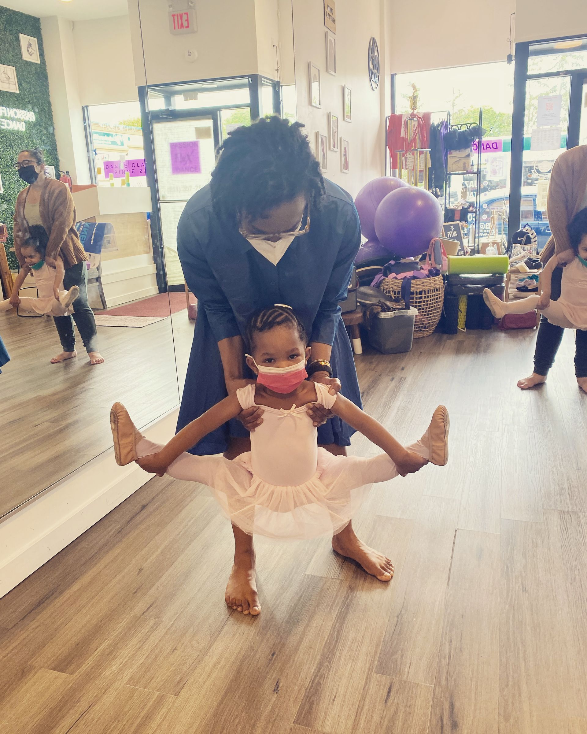 Instructor assisting young ballerina in a split; studio setting. They both wear masks.