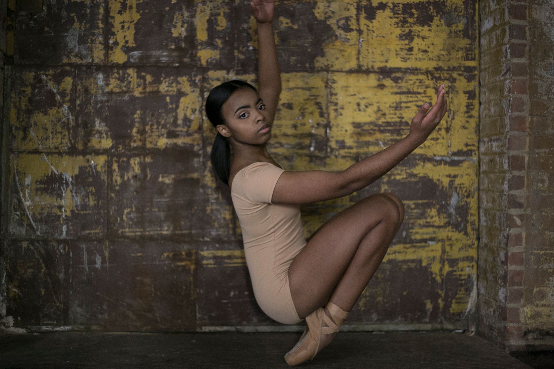 A Black dancer in a tan leotard and pointe shoes crouches, arms extended near a distressed yellow wall.