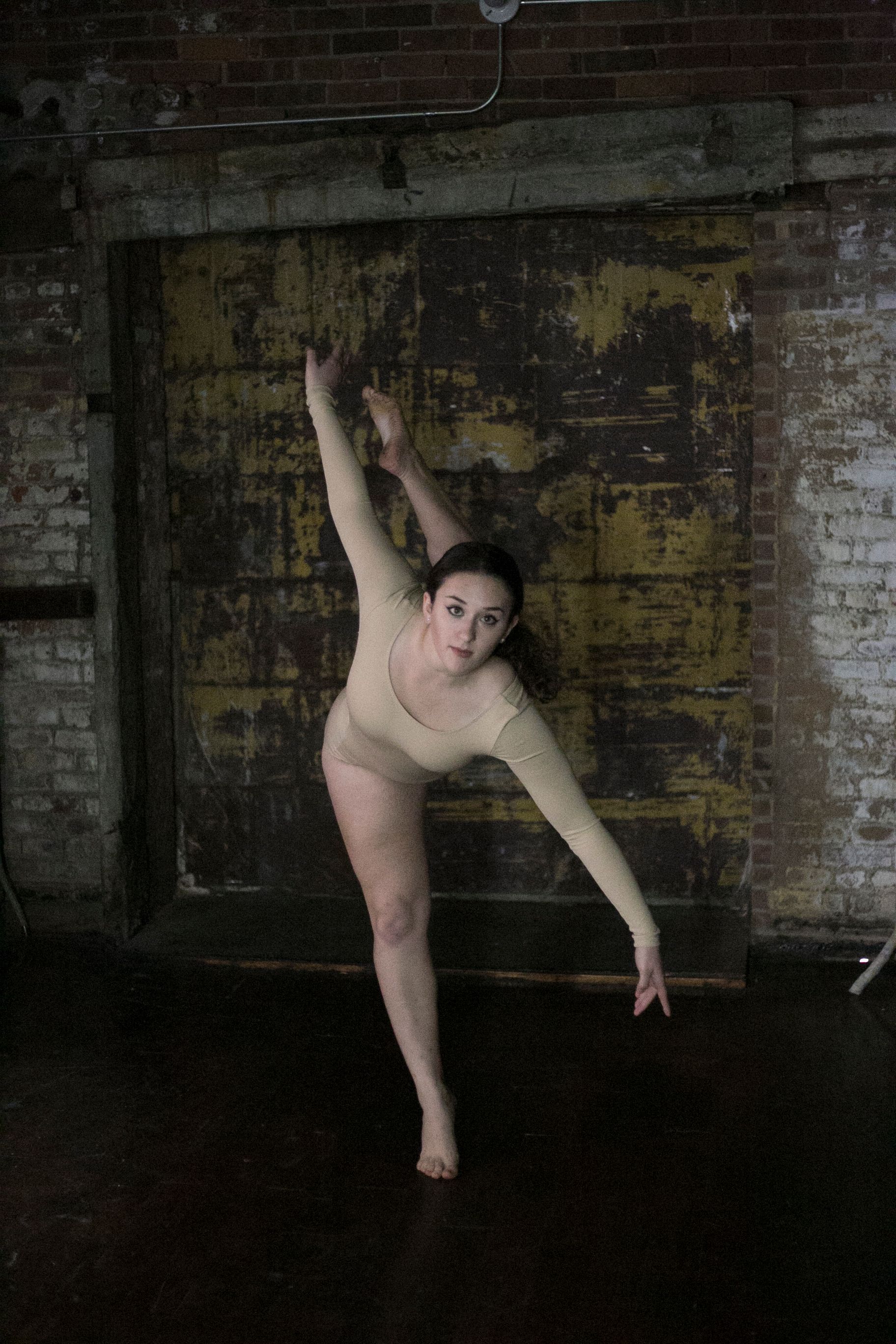 A dancer in a beige leotard performs a graceful pose against a distressed wooden door and brick wall.