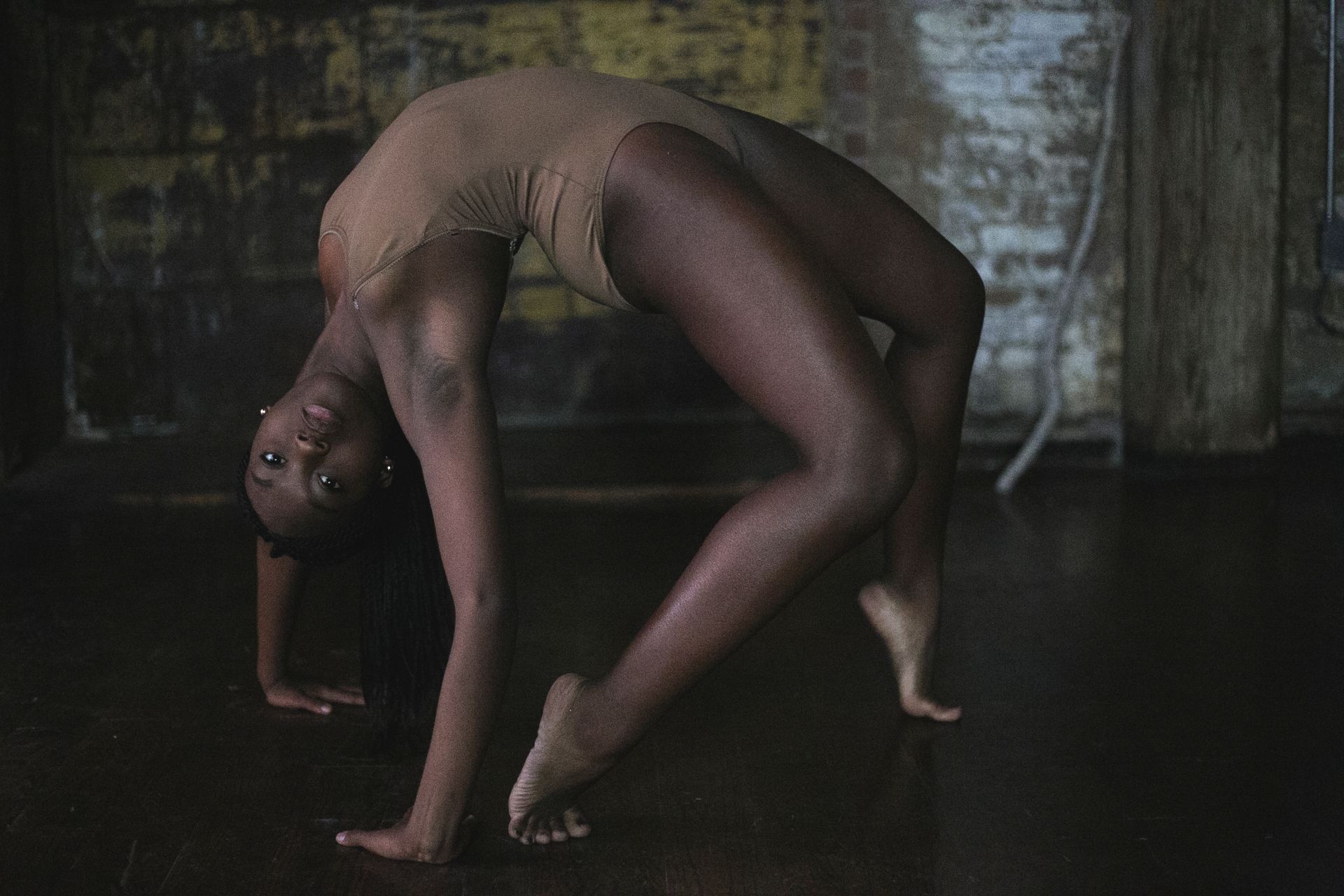 A dancer in a tan leotard arches backward in a dark, gritty space.