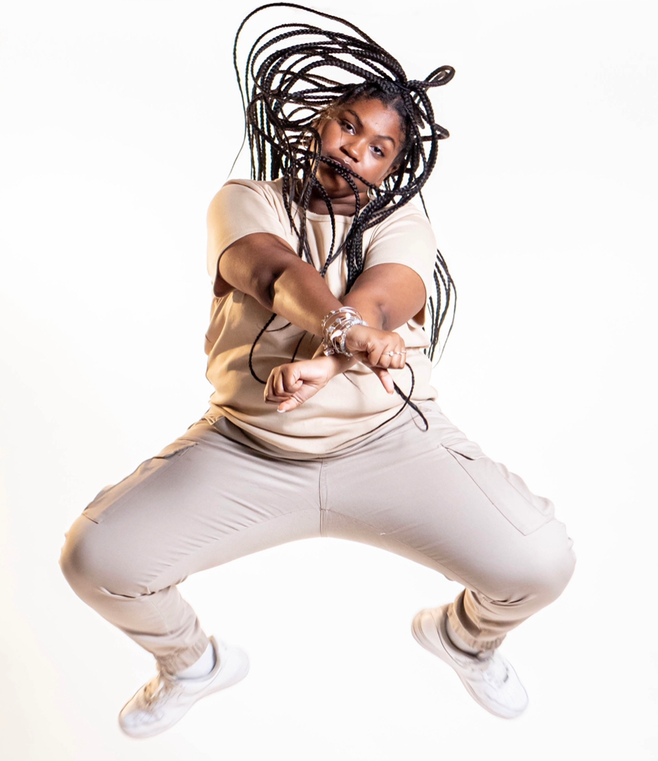 Black woman, jumping with long braids. She wears tan sweatsuit, white sneakers. White background.