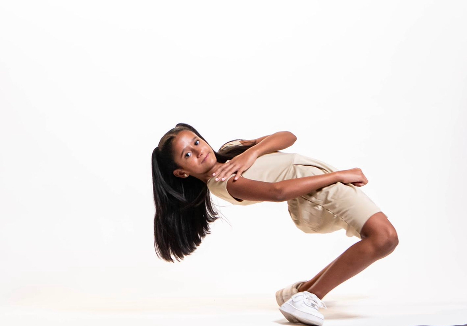 Young girl with long dark hair, bending backward in a dance pose, beige outfit, white background.