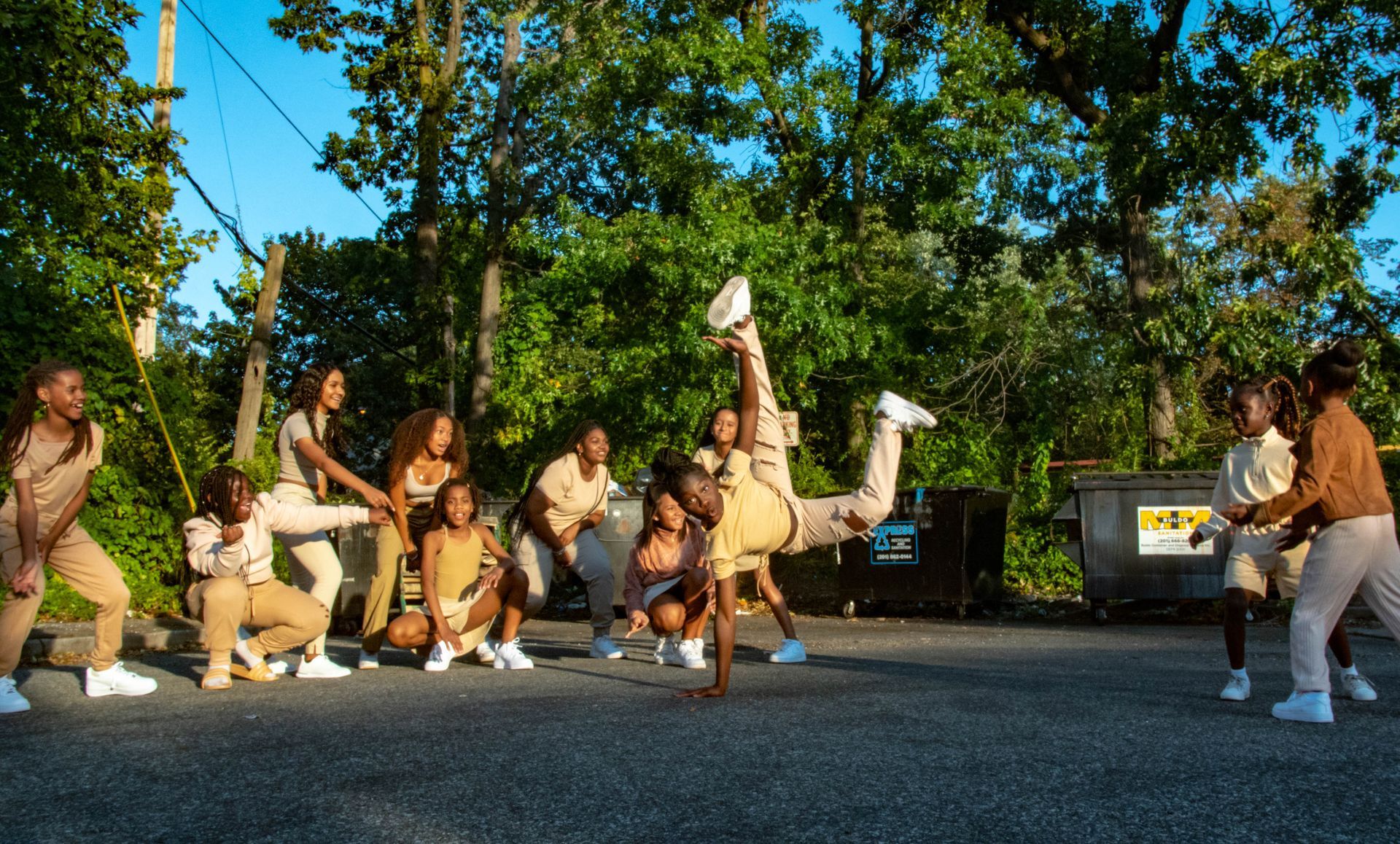 A group of people in tan outfits dance on pavement. One person does a handstand. Trees in the background.
