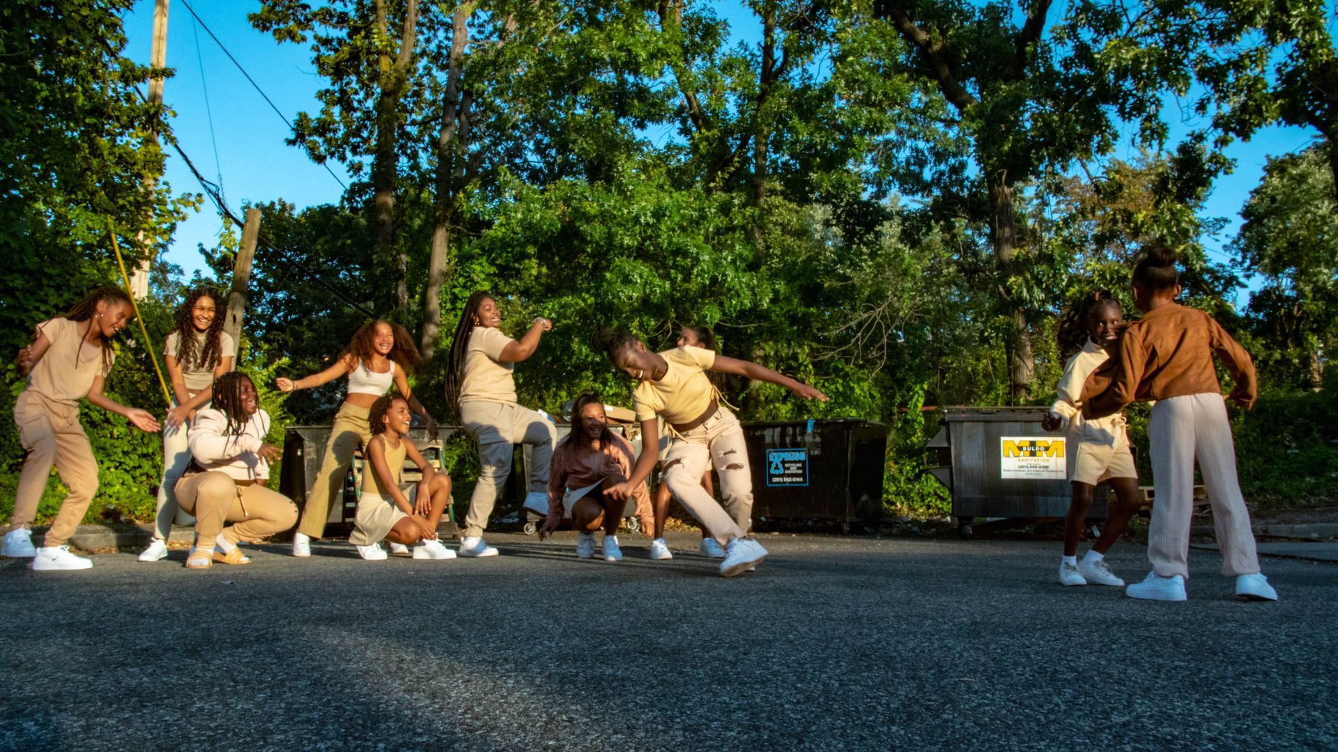 Group of dancers in beige outfits posing on a street, under trees, with a dumpster.