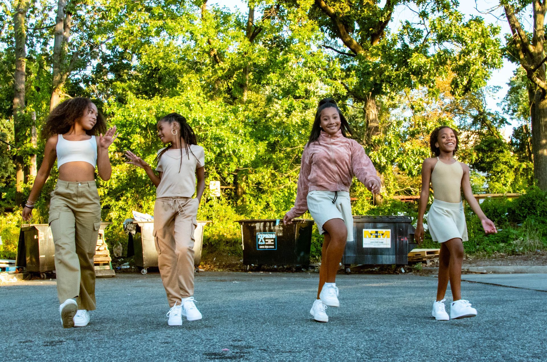 Four girls dance joyfully on an asphalt road, sunny, green trees in background. They are wearing casual, stylish clothes.