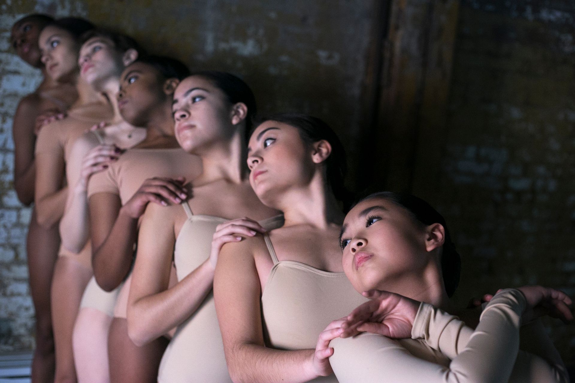 Eight ballet dancers in beige leotards, arms on each other's shoulders, looking up in a brick-walled space.