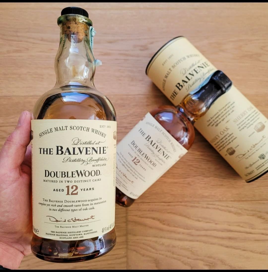 Una persona tiene in mano una bottiglia di whisky Balvenie Doublewood 12 anni. Un'altra bottiglia e un tubetto sono accanto, su una superficie di legno.