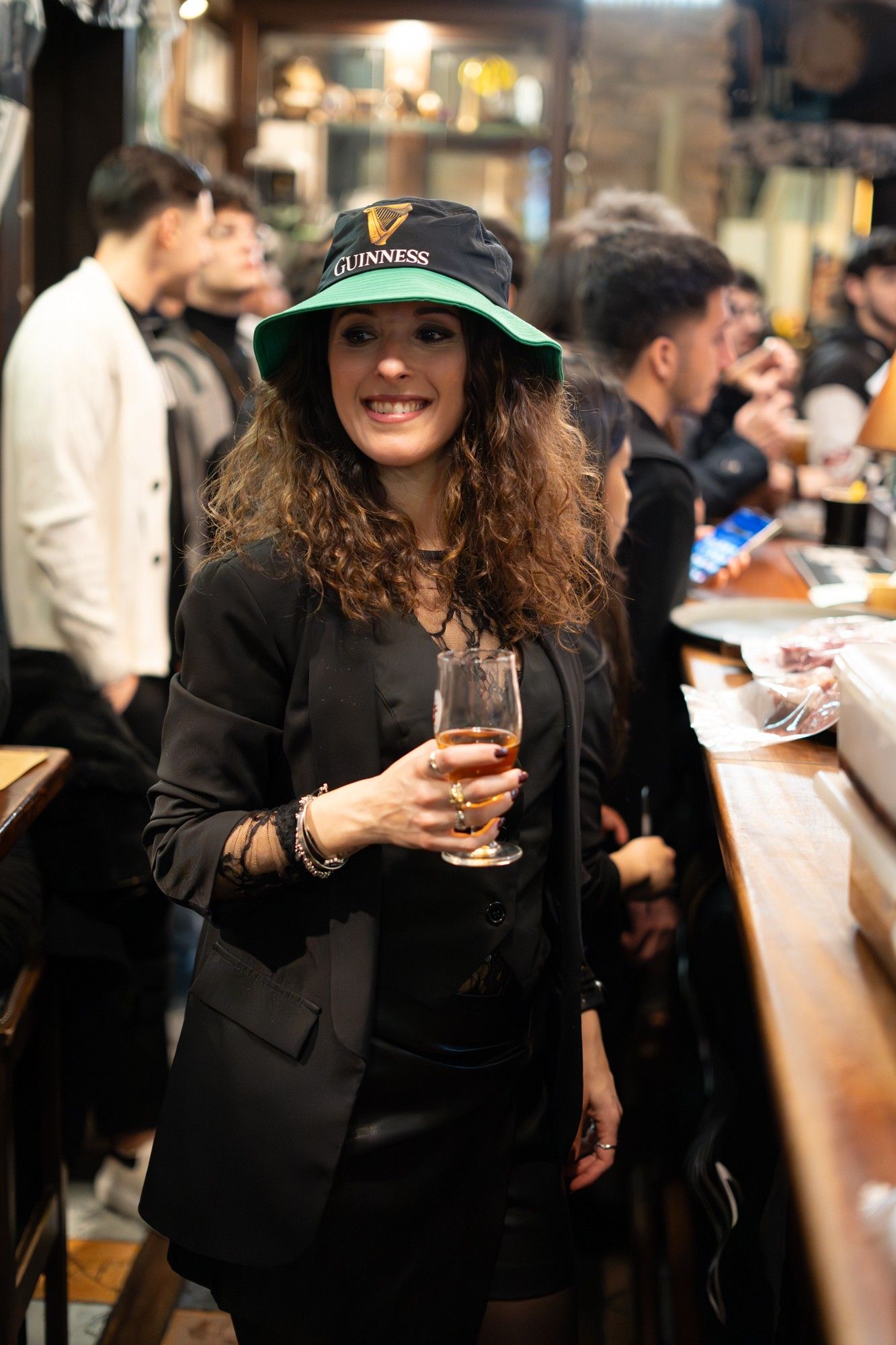 Donna in abito nero, cappello da pescatore Guinness, con in mano un drink al bar, sorridente.