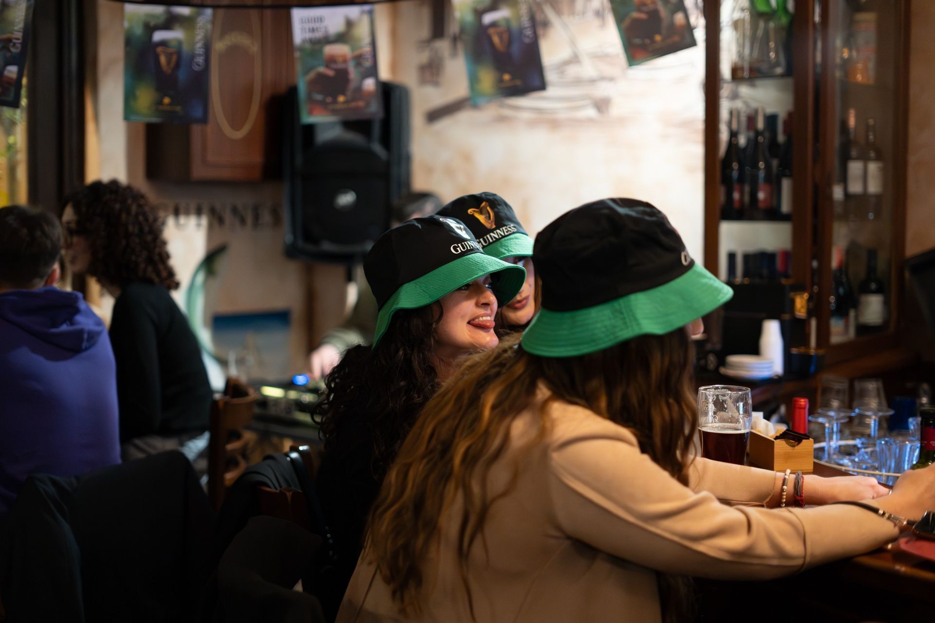 Due donne con cappelli verdi chiacchierano in un bar. Sullo sfondo, un poster della Guinness e bottiglie di alcolici.