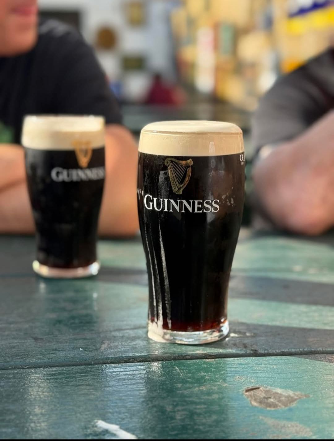 Due pinte di Guinness stout su un tavolo verde con lo sfondo sfocato di un pub, una delle quali è a fuoco.