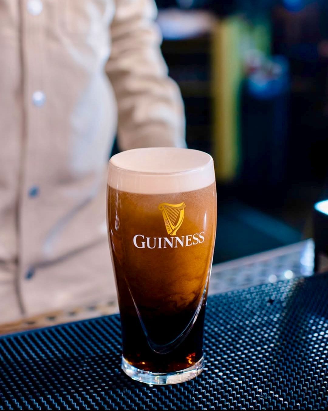 Una pinta di Guinness con una schiuma cremosa, appoggiata su un bancone con un barista sullo sfondo.