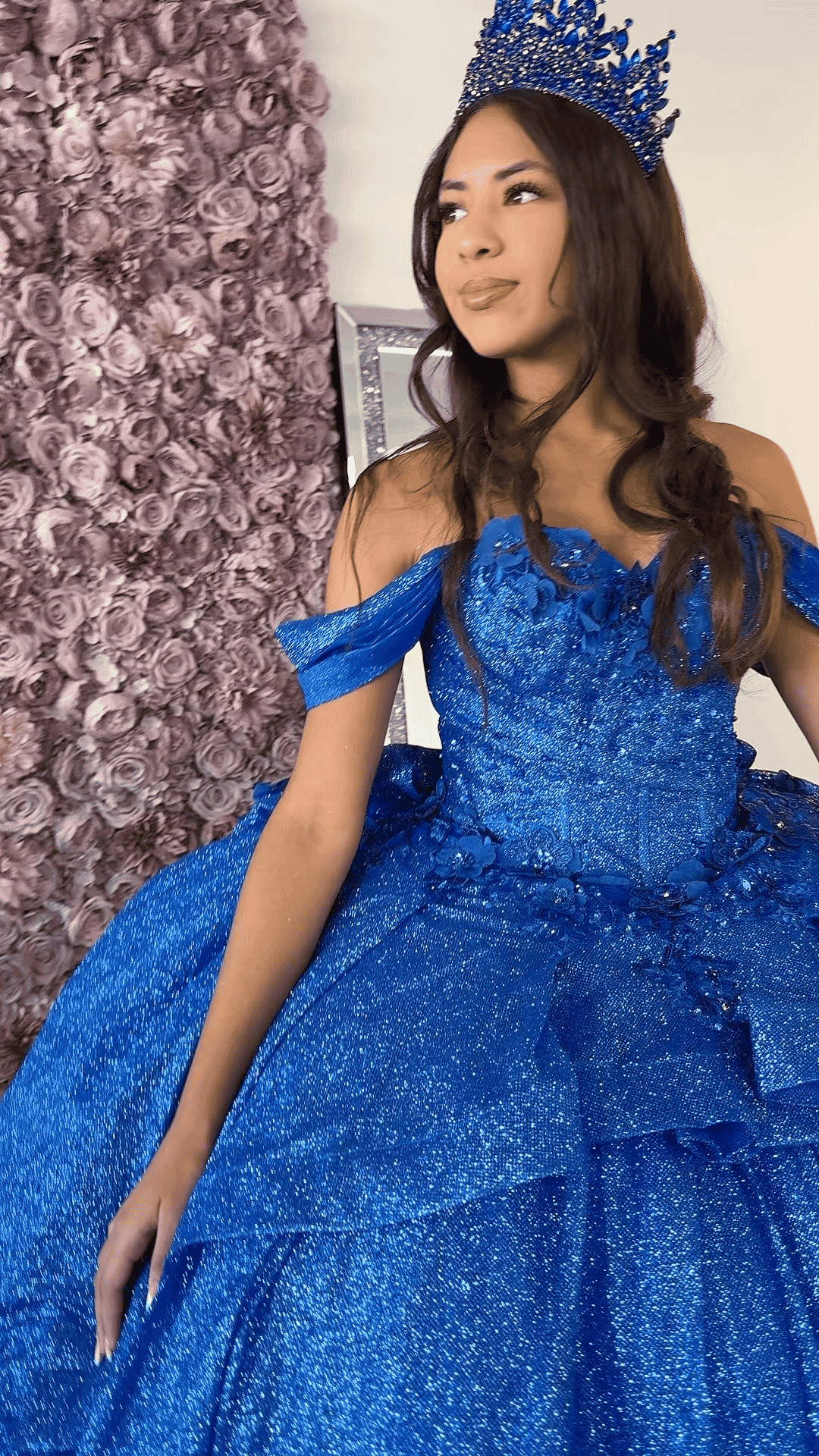 A woman in a blue dress and crown is standing in front of a wall of flowers.