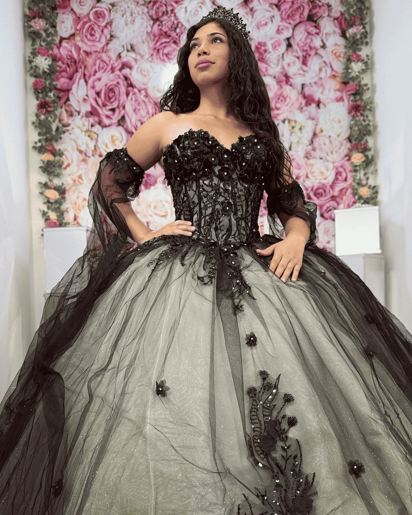 A woman in a black and white ball gown is standing in front of a wall of flowers.