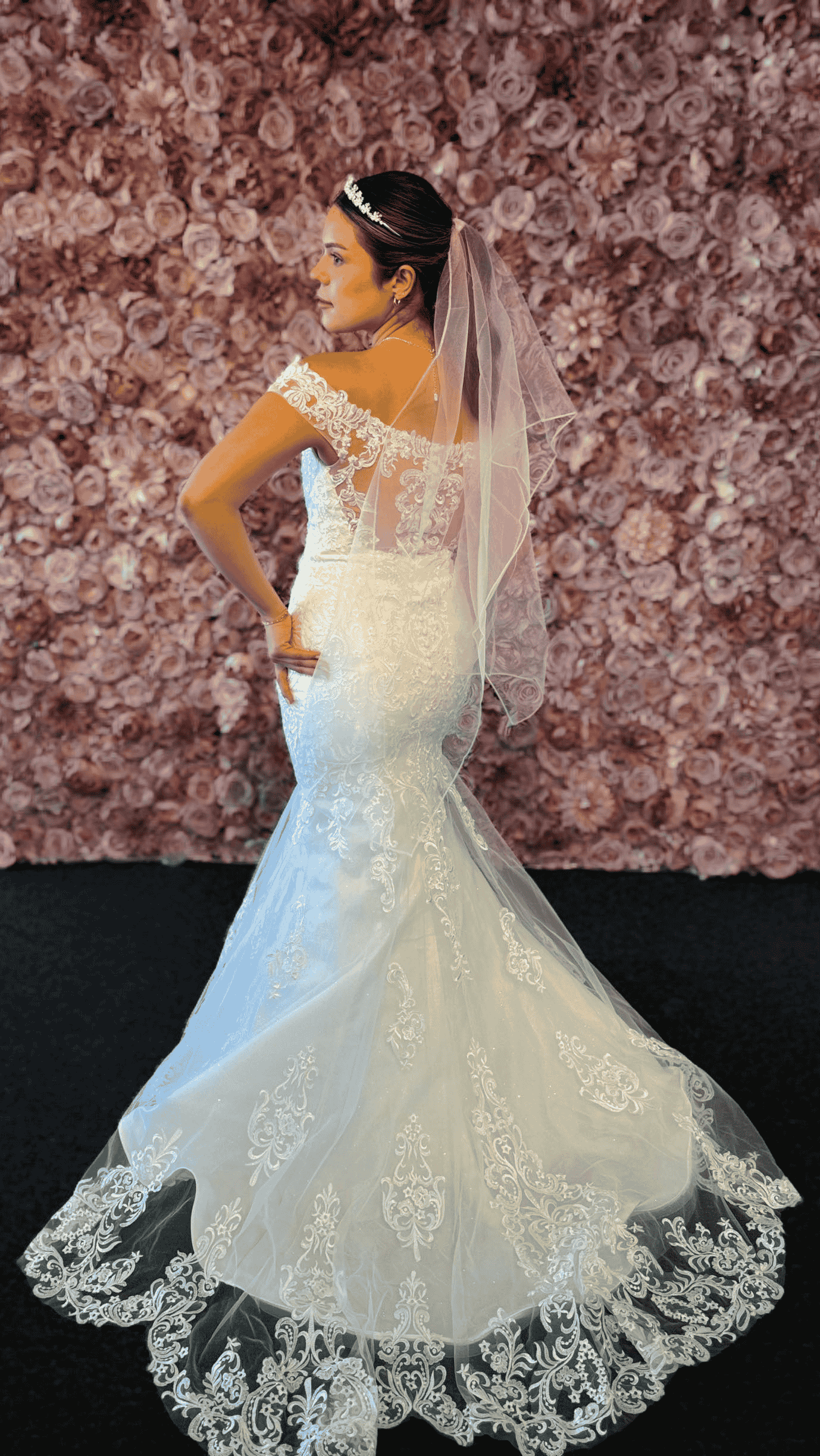 A woman in a wedding dress and veil is standing in front of a wall of flowers.