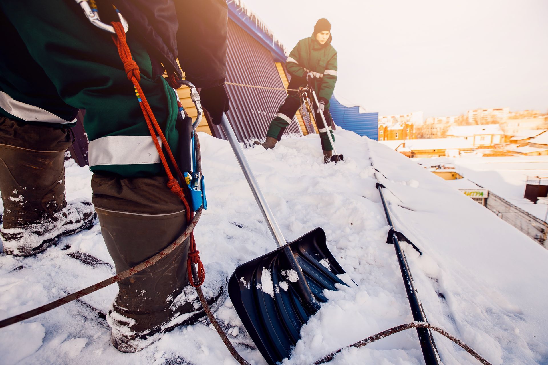 Workers shoveling snow off a rooftop, blue sky, one person roped in, using black shovels, winter.
