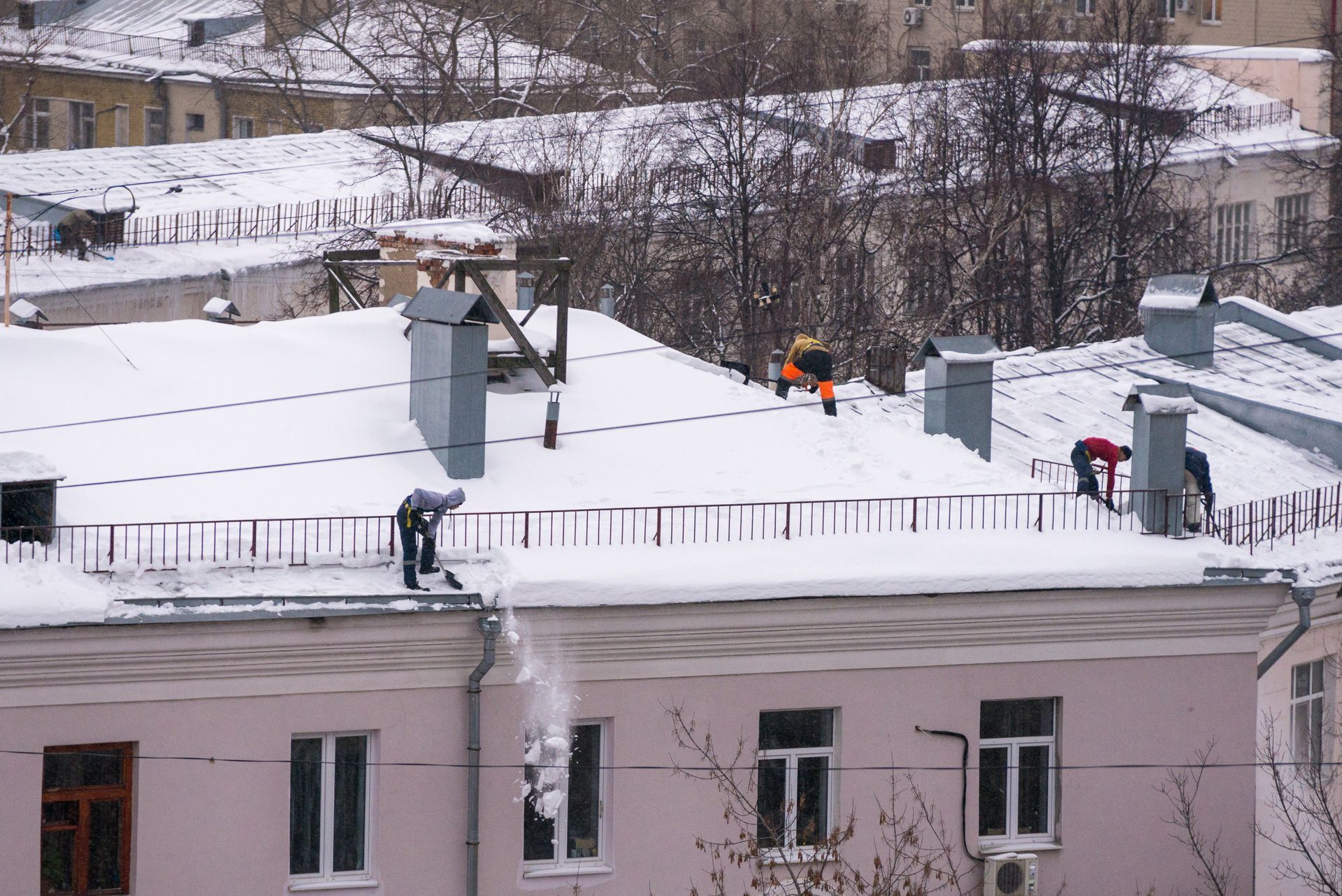 Workers clearing snow from a building's roof in a wintry urban setting.