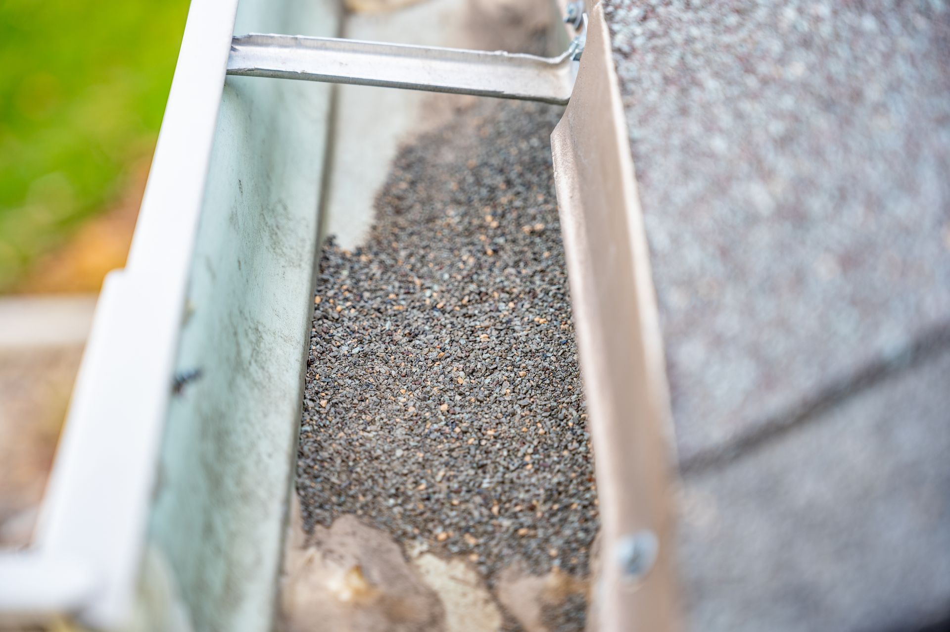 Close-up of a gutter filled with dark debris and granules; part of a house roof is visible.