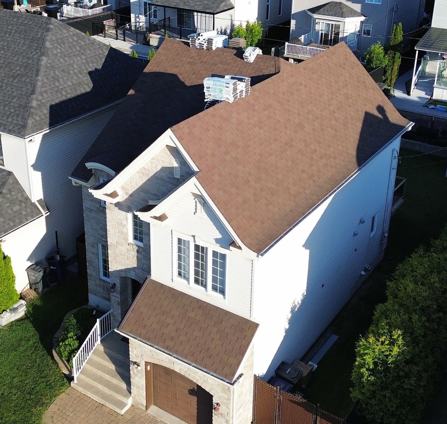 Vue plongeante d'une maison au toit brun, à la façade en briques beiges et au garage double. La lumière du soleil projette des ombres sur l'extérieur.