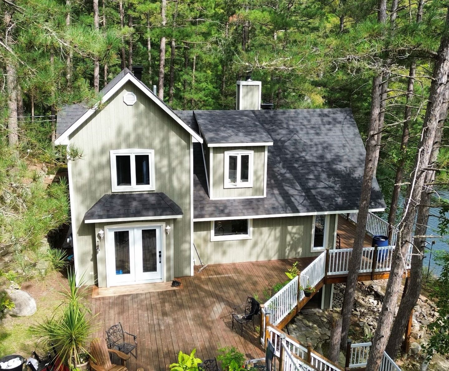 Une maison verte à deux étages avec un toit noir se trouve sur une terrasse en bois entourée d'arbres, avec un lac visible en arrière-plan.