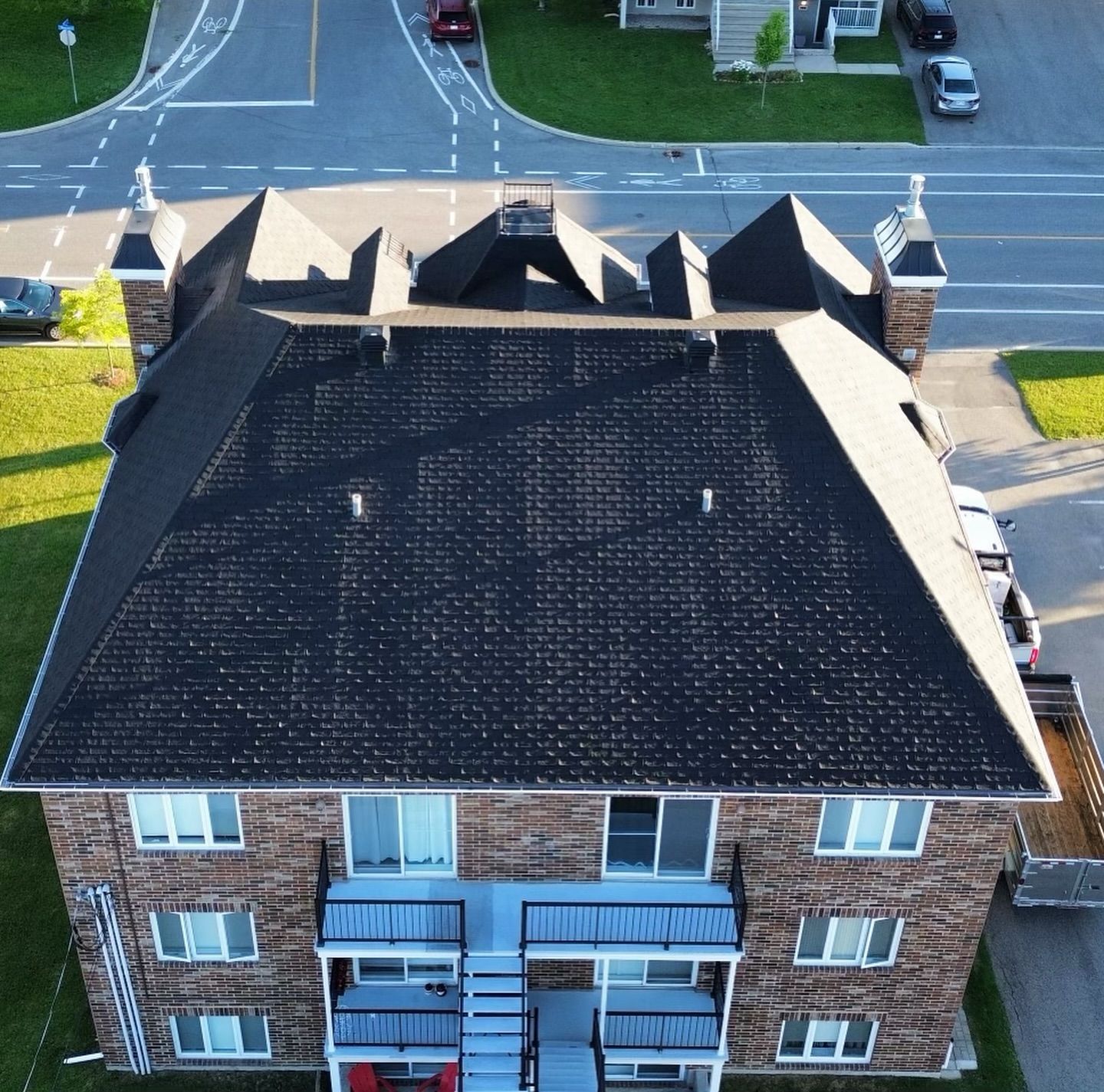 Vue aérienne d'un immeuble en briques avec un toit en bardeaux foncés. L'herbe verte entoure le bâtiment, laissant apparaître une rue et des voitures garées.