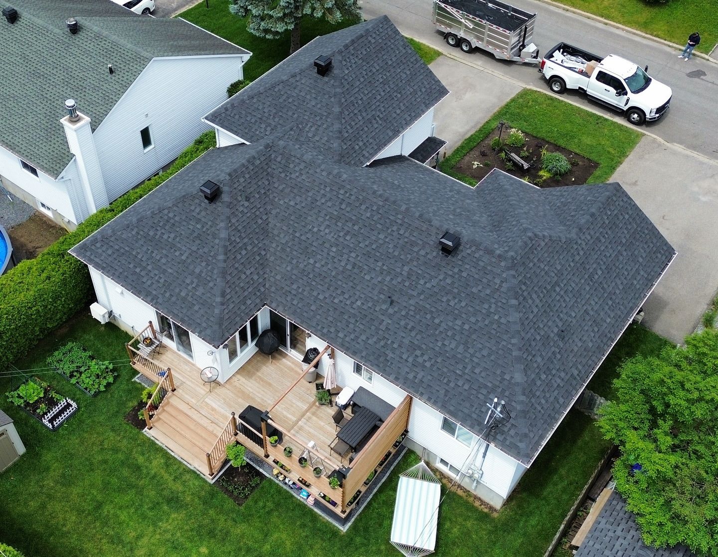 Vue aérienne d'une maison avec un toit gris foncé nouvellement recouvert de bardeaux, un revêtement blanc et une terrasse en bois dans un cadre suburbain.