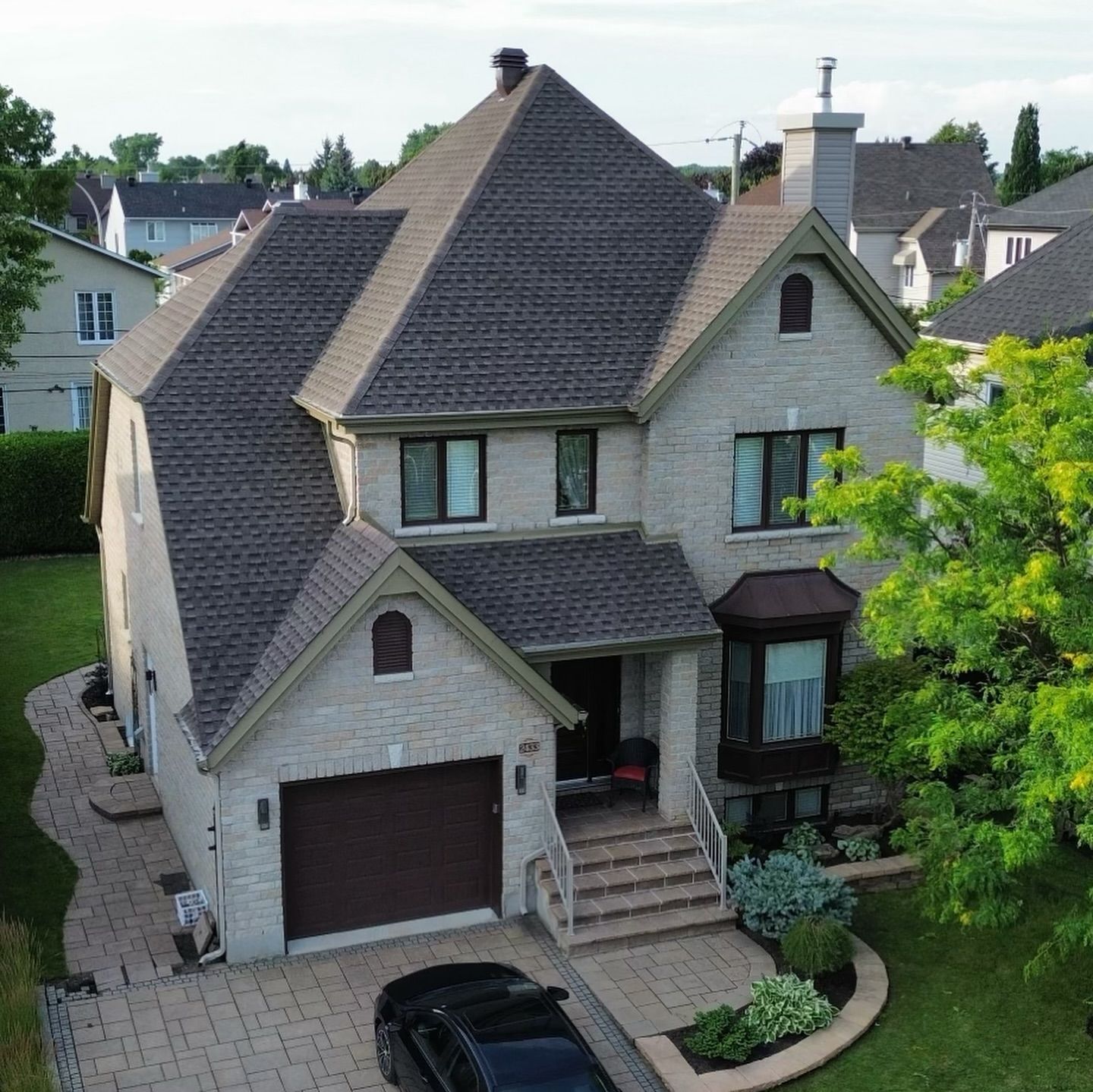 Une maison en briques à deux étages avec un toit brun foncé, une allée et un garage attenant. Une voiture noire est garée dans l'allée.