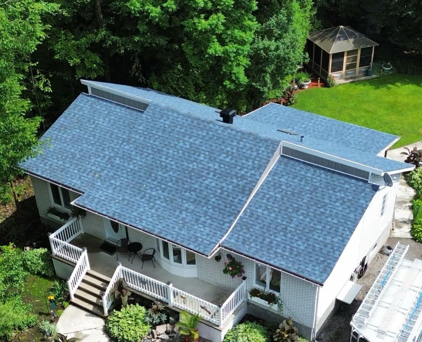 Vue aérienne d'une maison blanche avec un toit en bardeaux bleus, entourée d'arbres verts et d'un belvédère dans l'arrière-cour.