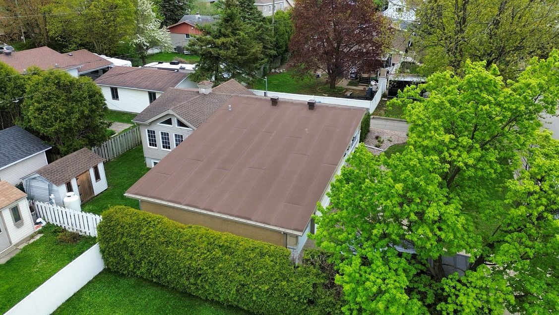 Vue aérienne d'une maison au toit brun, avec une cour verte et les maisons environnantes. Un grand arbre vert vif est au premier plan.