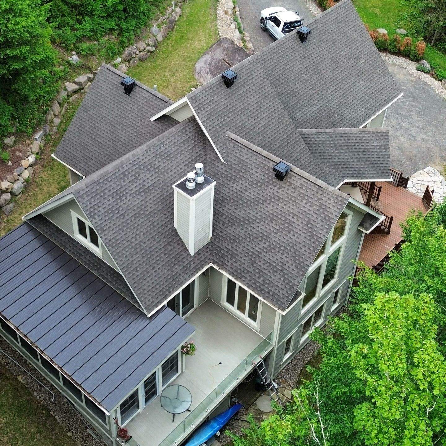 Vue aérienne d'une maison avec un toit gris foncé et un revêtement vert clair, entourée d'arbres et d'une allée avec une voiture blanche.