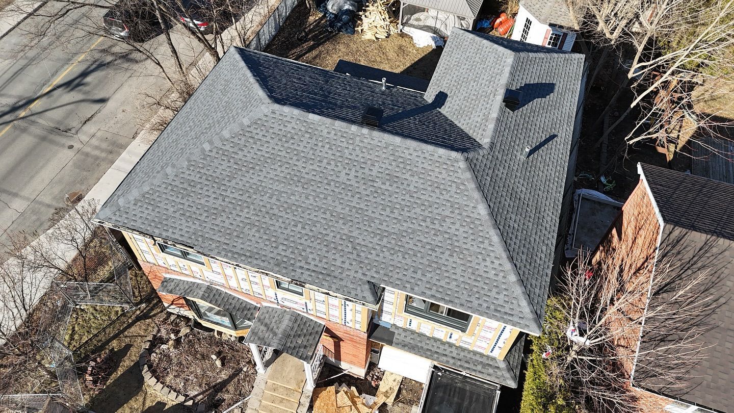 Vue aérienne d'une maison à deux étages avec un toit en bardeaux gris. La maison possède une façade en briques et est entourée d'arbres.
