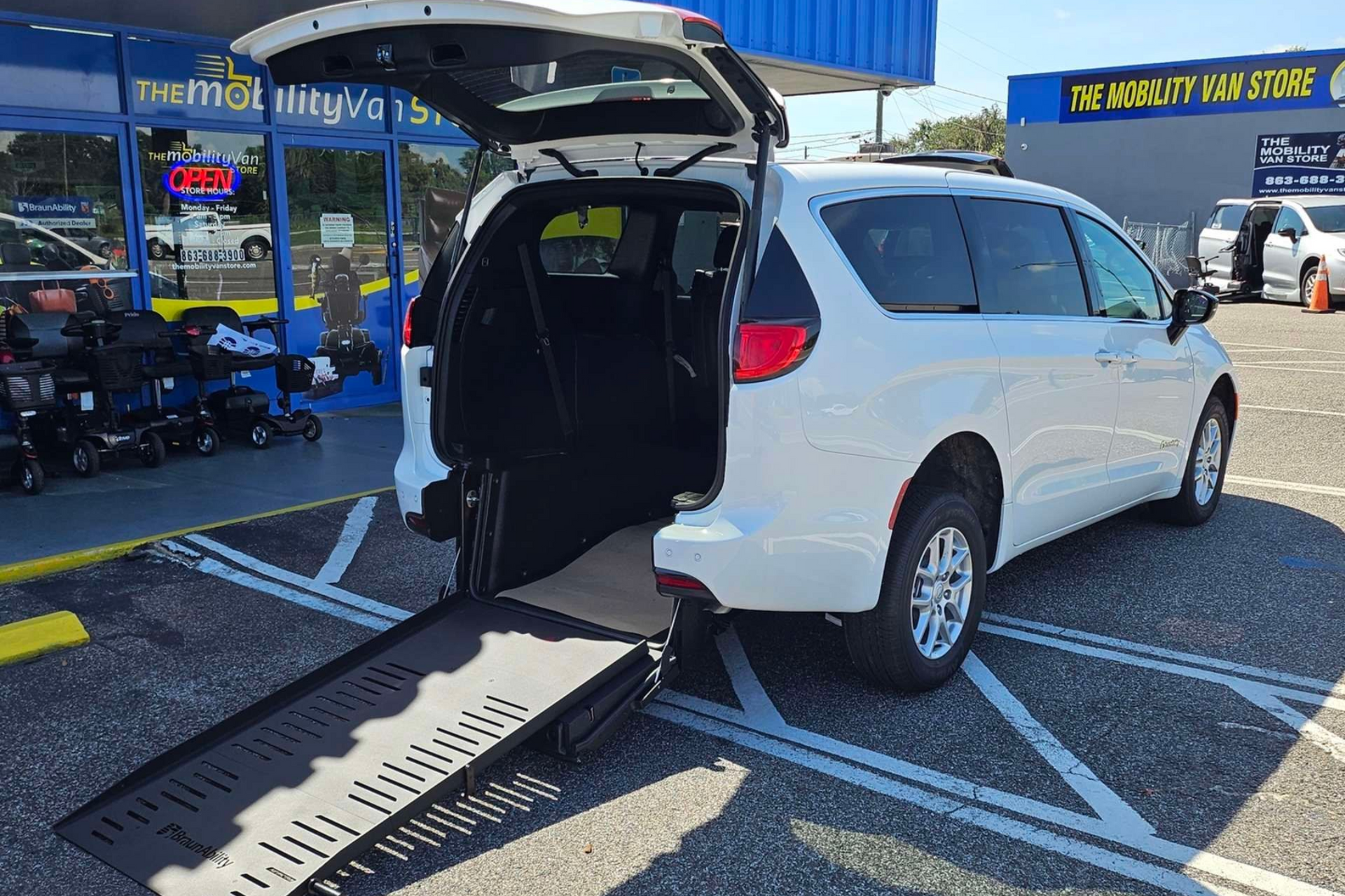 White wheelchair accessible van with ramp extended, parked at a store with mobility aids.
