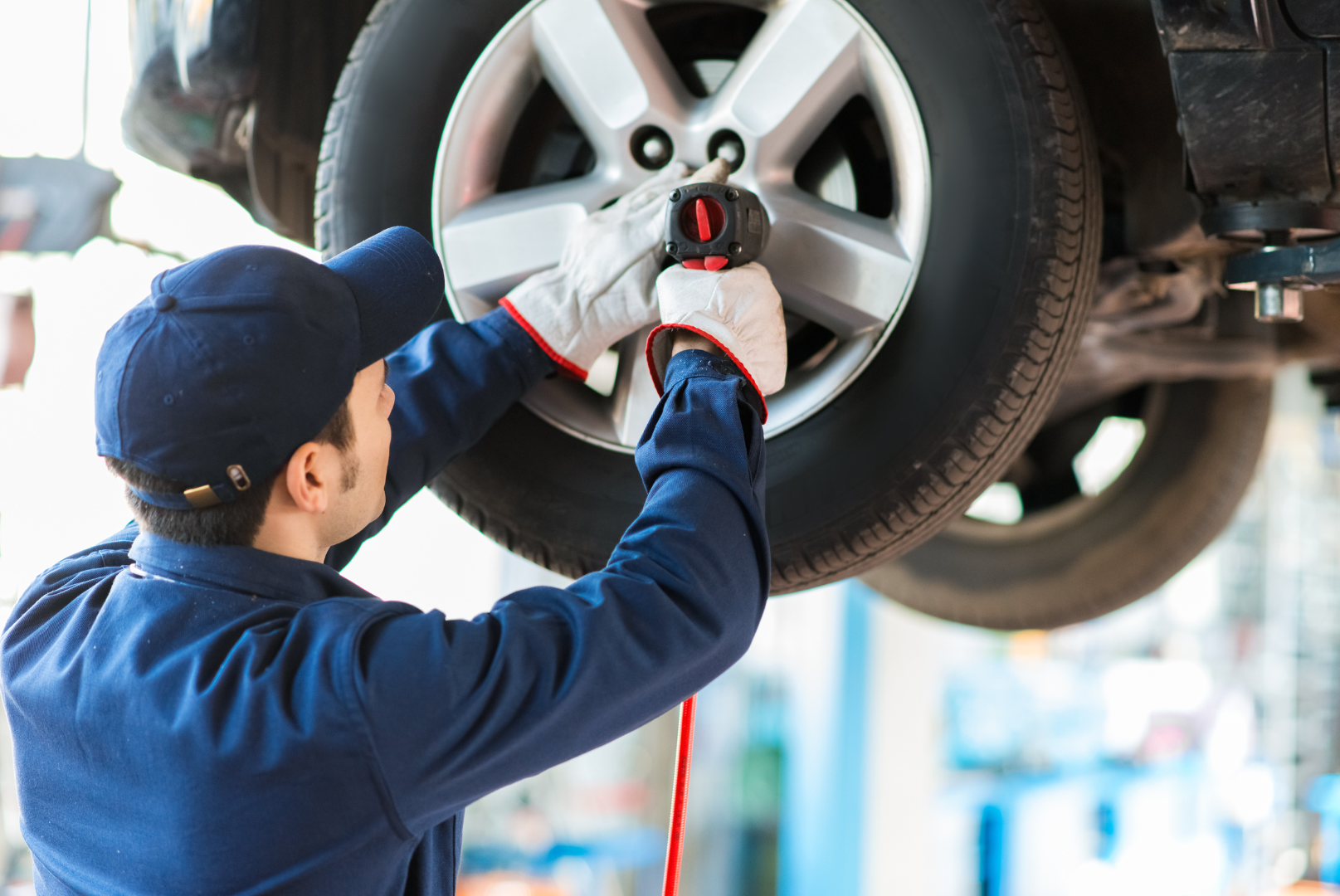 Mechanic in blue jumpsuit removing a car tire with an electric wrench in a garage.