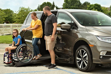 Family helping a girl in a wheelchair into a modified van. Person in yellow shirt and man help with the ramp. Outdoors.