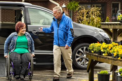 Woman in wheelchair smiles at man opening accessible van door at a plant nursery.