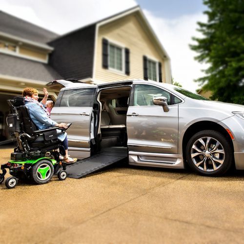 Person in wheelchair exiting a silver accessible van with a ramp in front of a house.