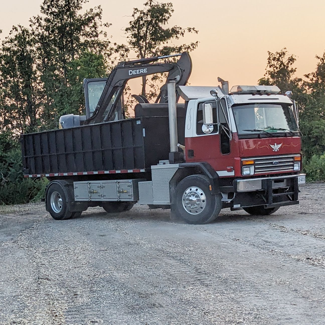 A red and white commercial dump truck carrying a piece of heavy construction equipment on a gravel lot at dusk.