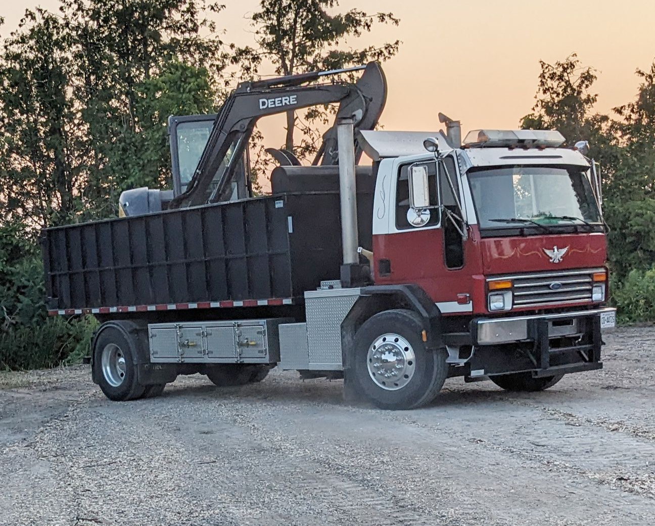 A red and white flatbed truck parked on gravel, carrying a heavy excavator machine in its cargo bed.