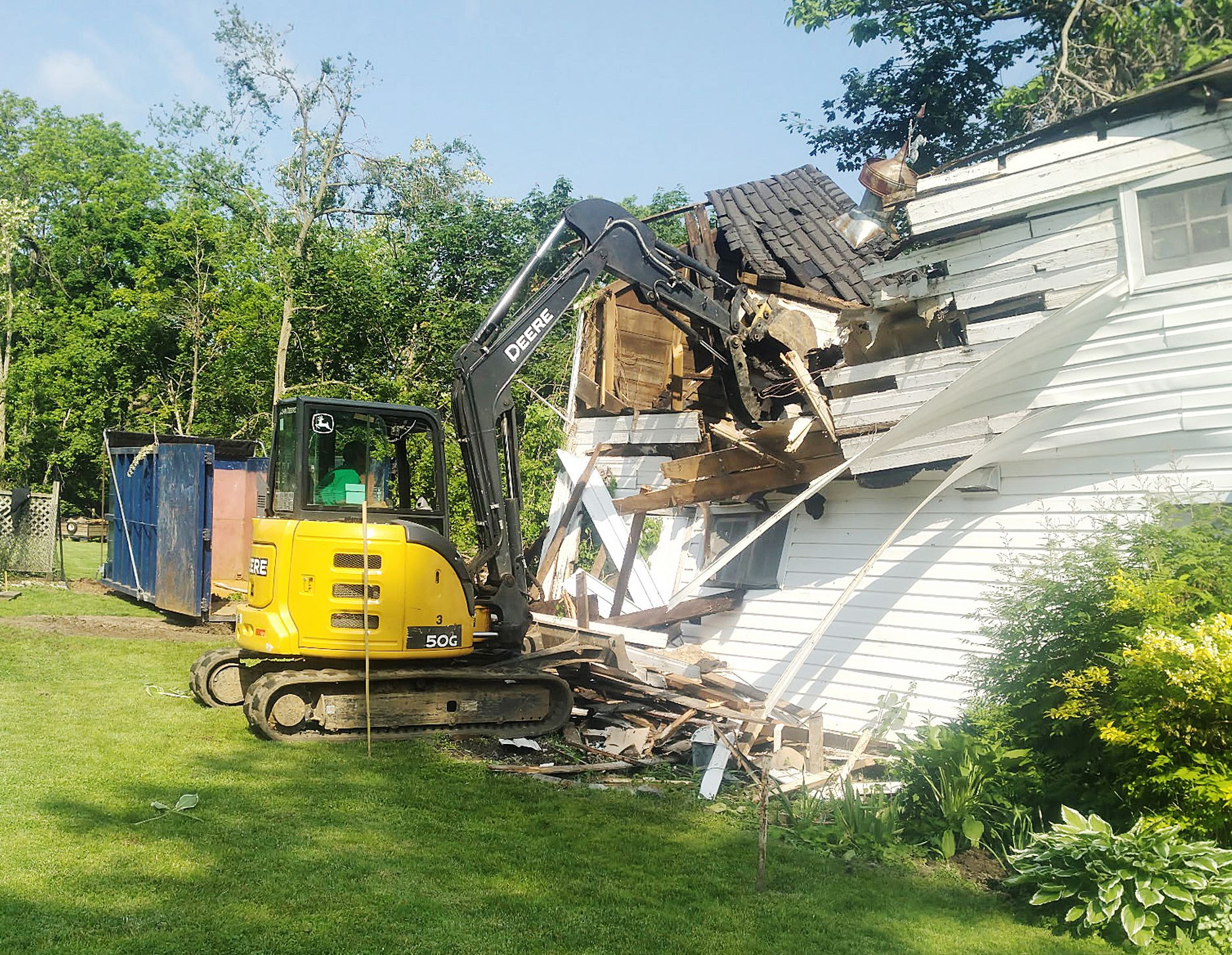 A yellow John Deere excavator tears down a section of a white, wooden house in a grassy, sunlit yard.