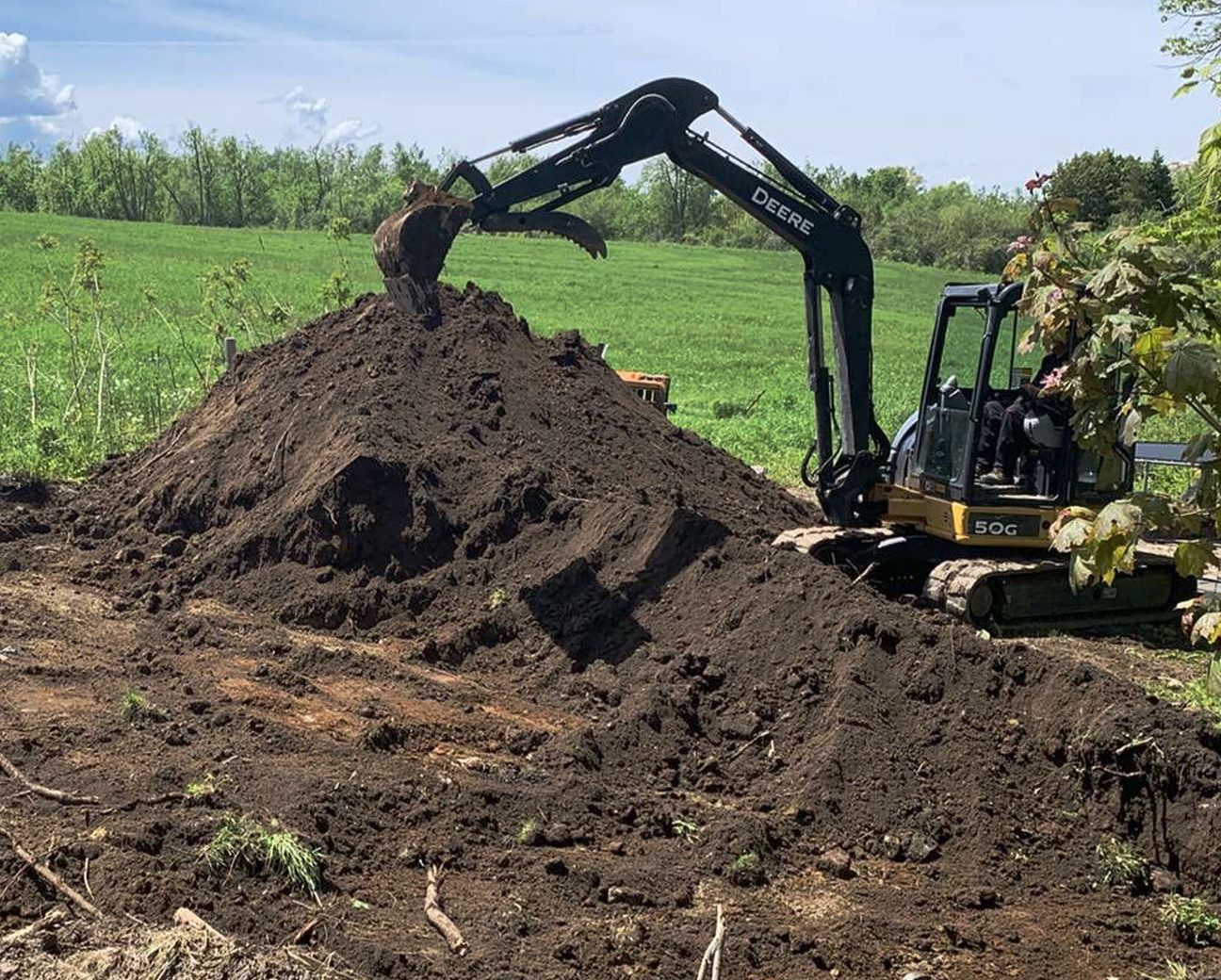 A black John Deere excavator moving a large pile of dark brown soil in a sunny, rural field.