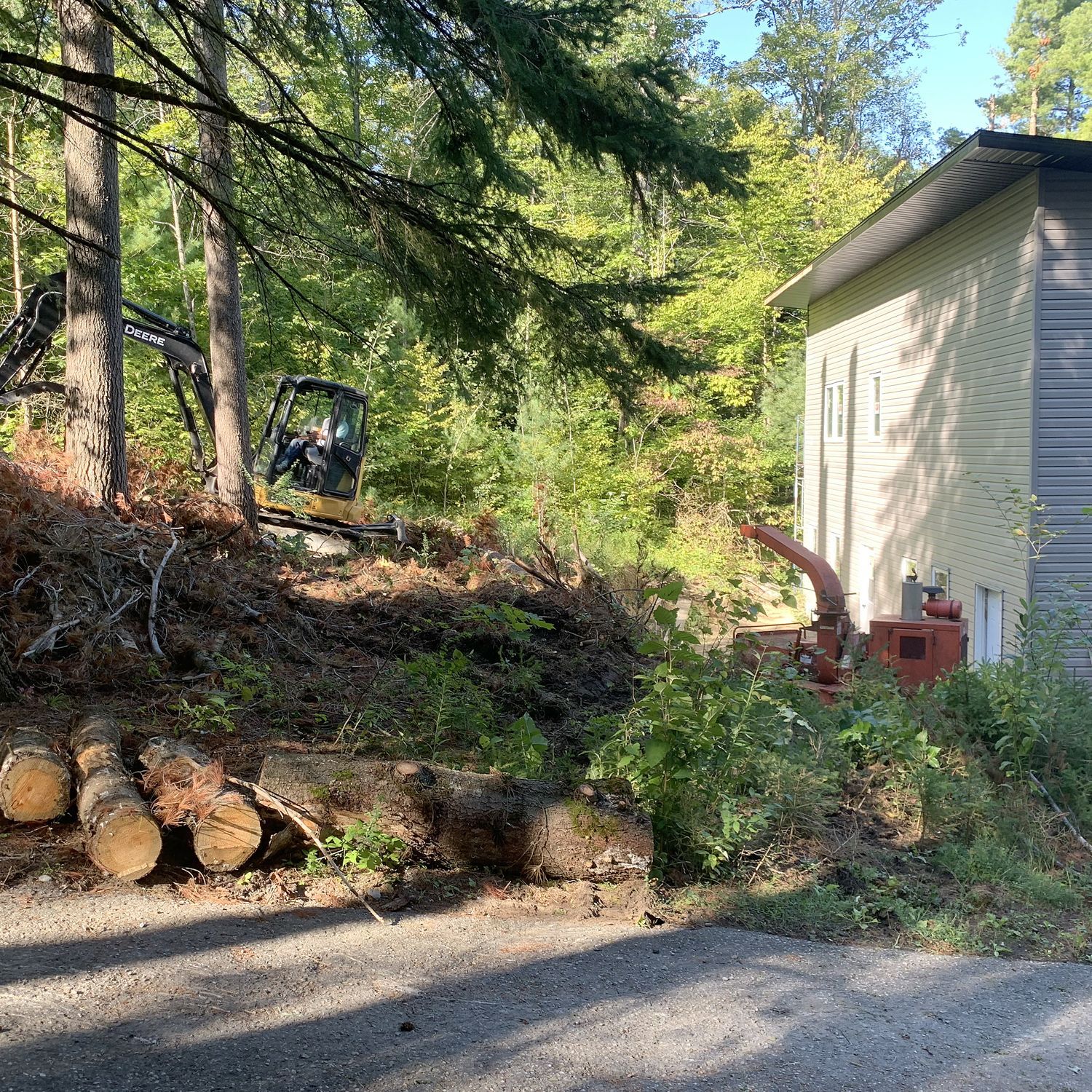 A construction excavator works in a wooded area next to a light-colored house, with cut logs in the foreground.