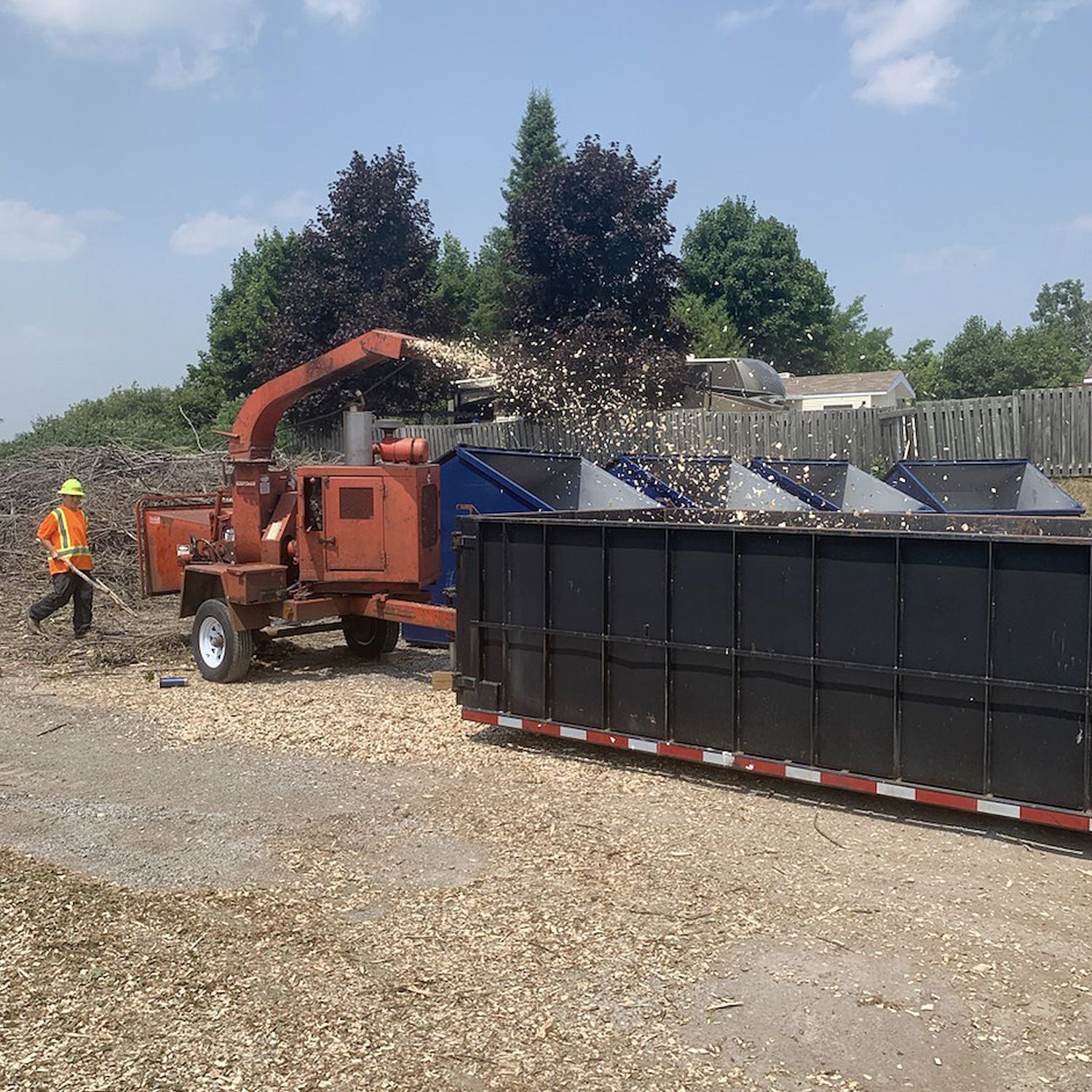 A worker in high-visibility gear operates a wood chipper, spraying wood chips into a large metal dumpster outdoors.