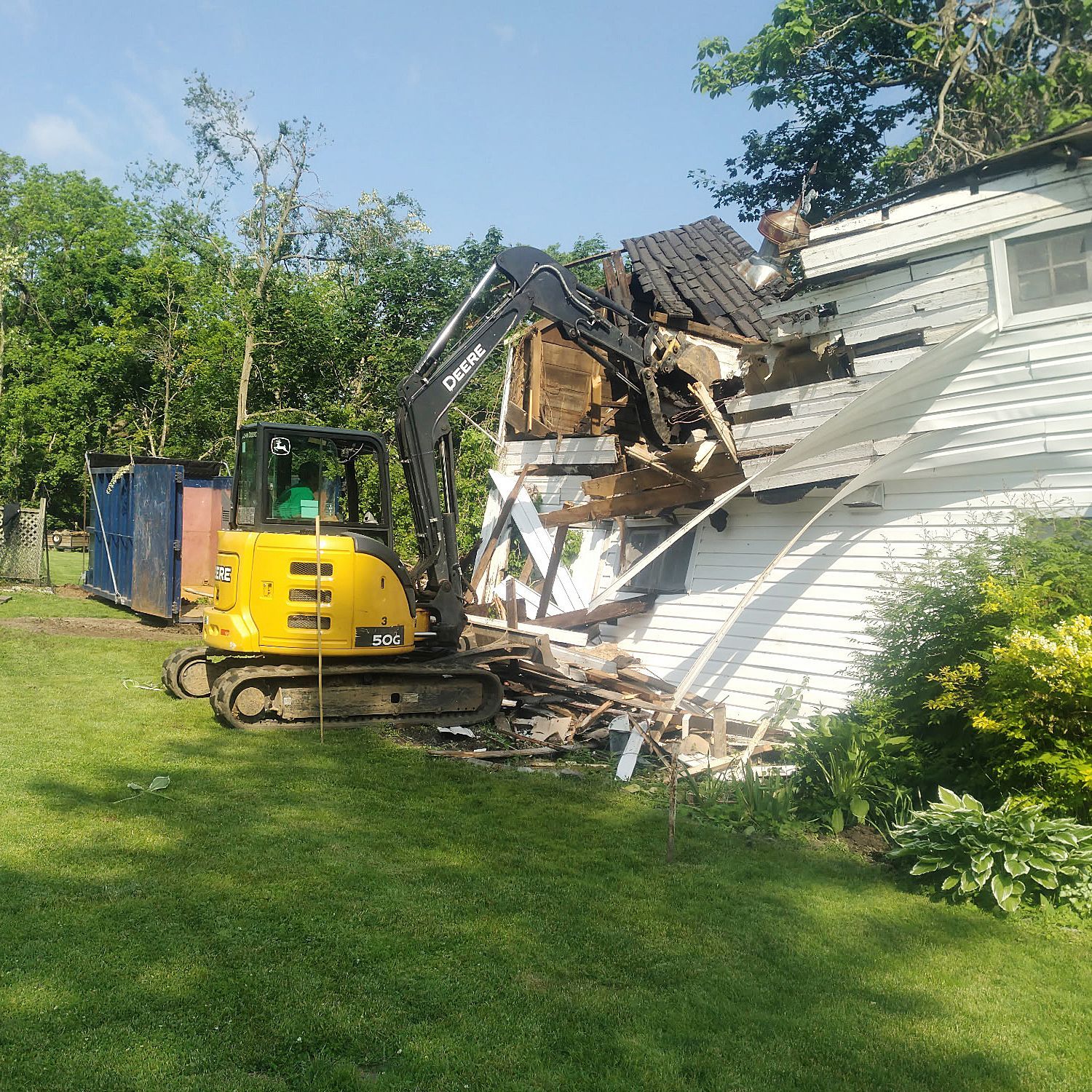 A yellow excavator demolishes the side of a small white house in a grassy yard.