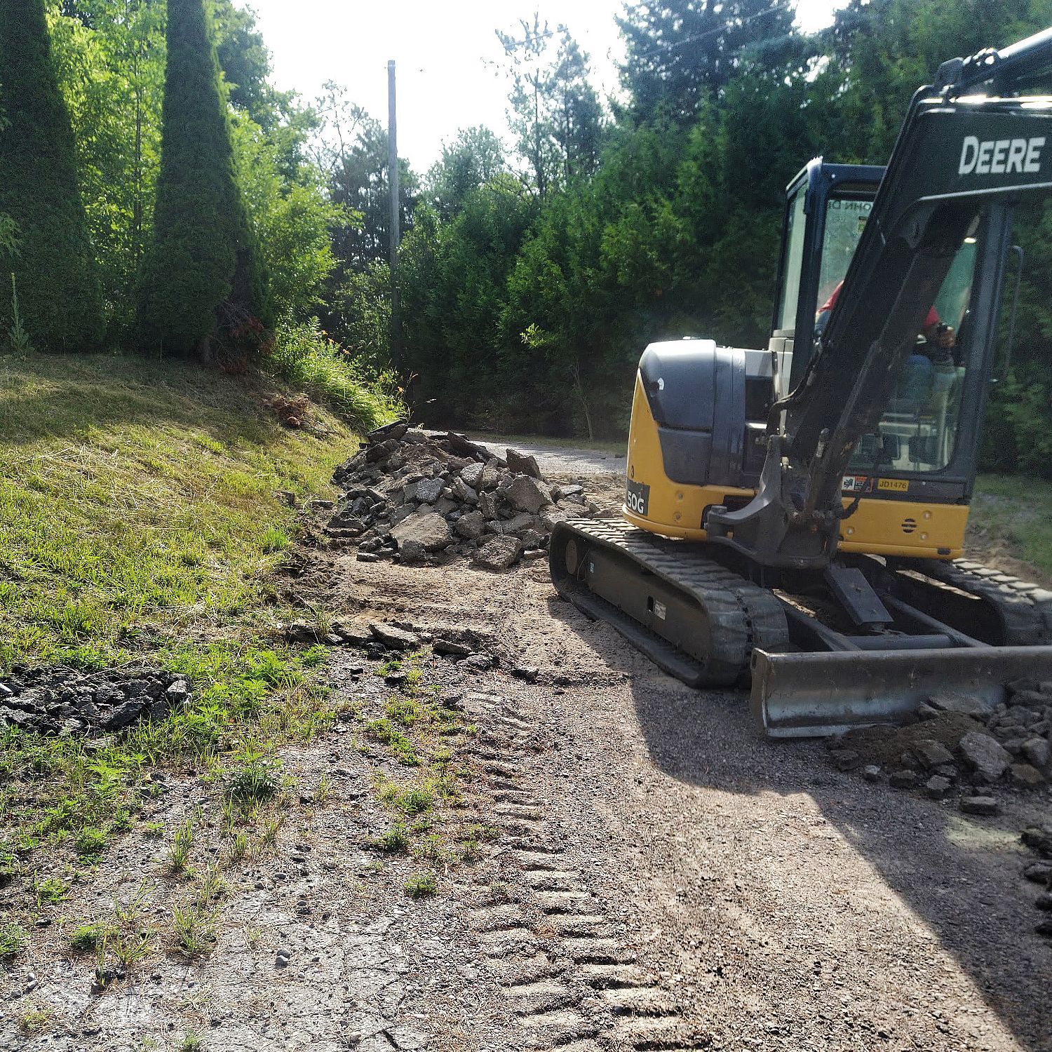 A John Deere excavator parked on a gravel path near a grassy slope and dense trees.