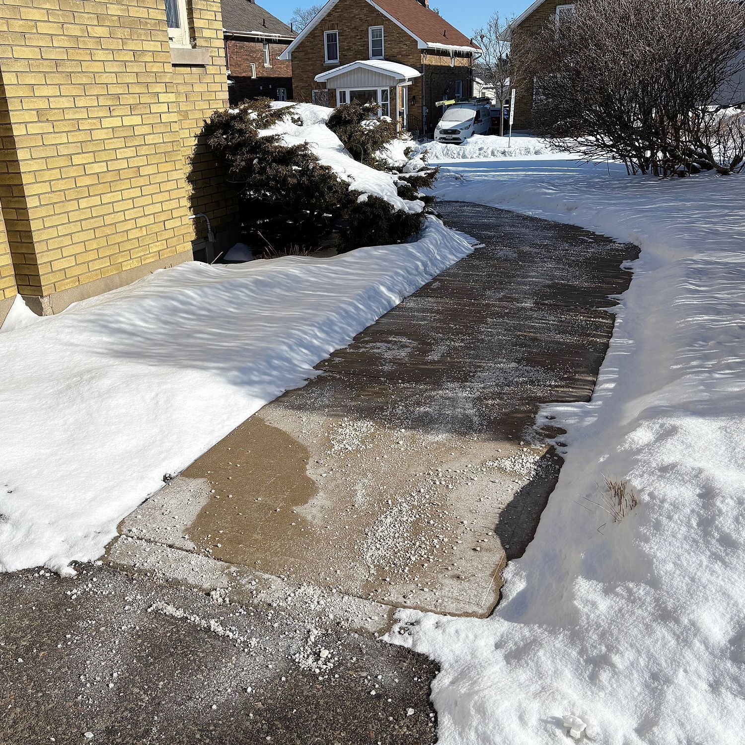 A concrete walkway leads to a house, flanked by piles of snow and a snow-covered bush on a bright winter day.