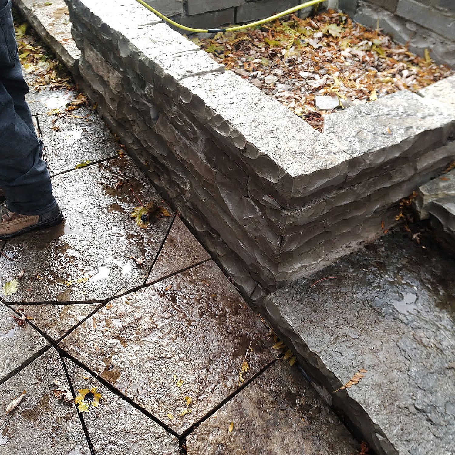 A person stands beside a low stone retaining wall and a flagstone patio covered with scattered autumn leaves.
