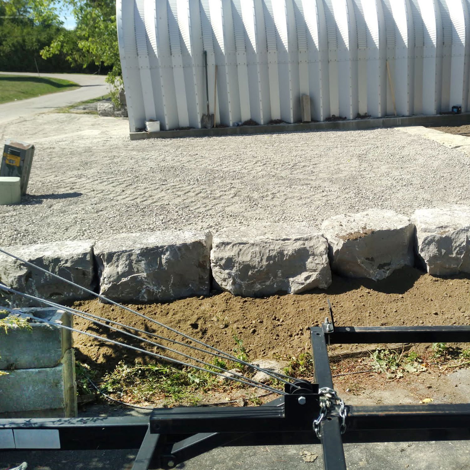 A metal frame with chains in the foreground sits before a row of large stone blocks and a white corrugated metal building.