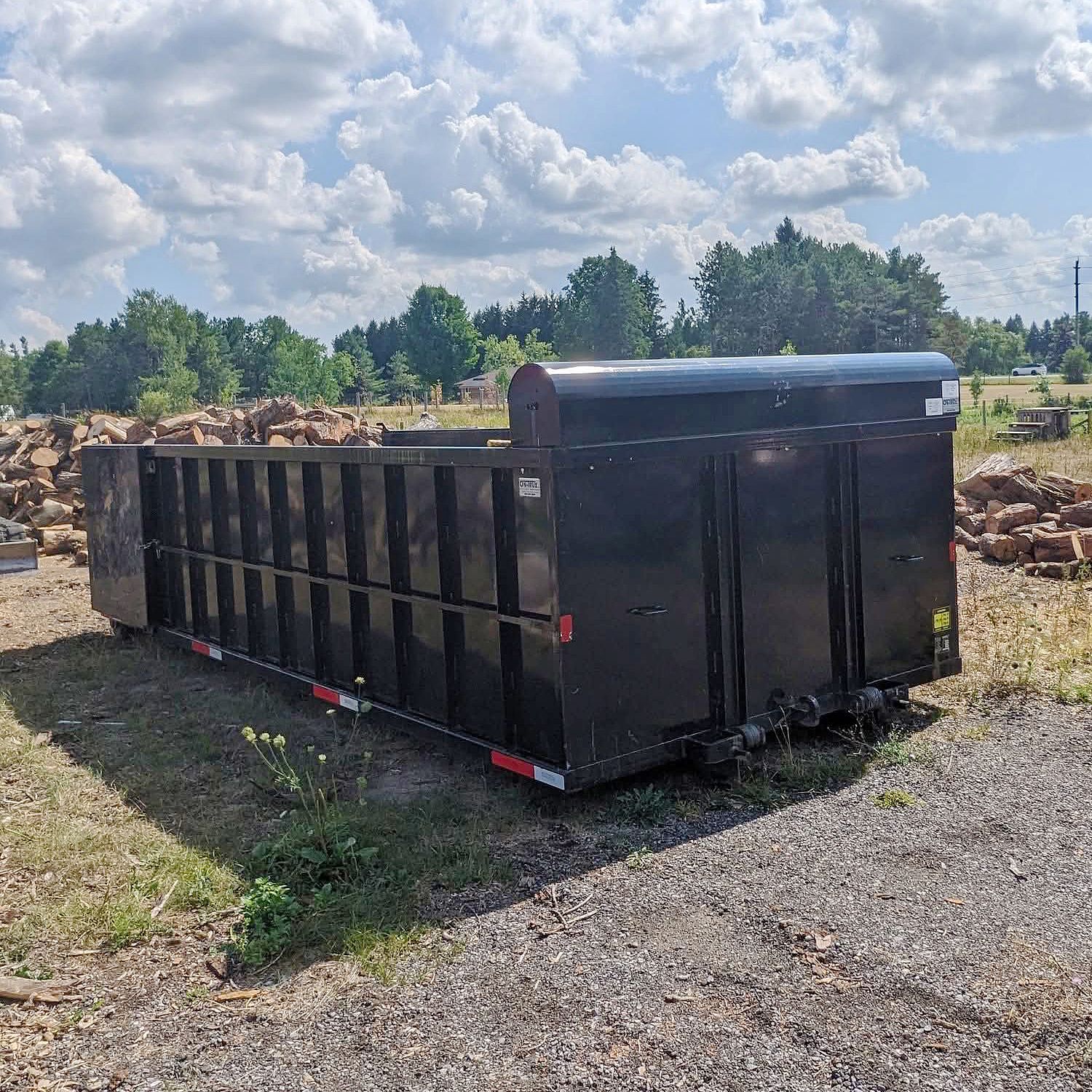 A large, black rectangular metal industrial dumpster sitting on a gravel lot under a partly cloudy sky.