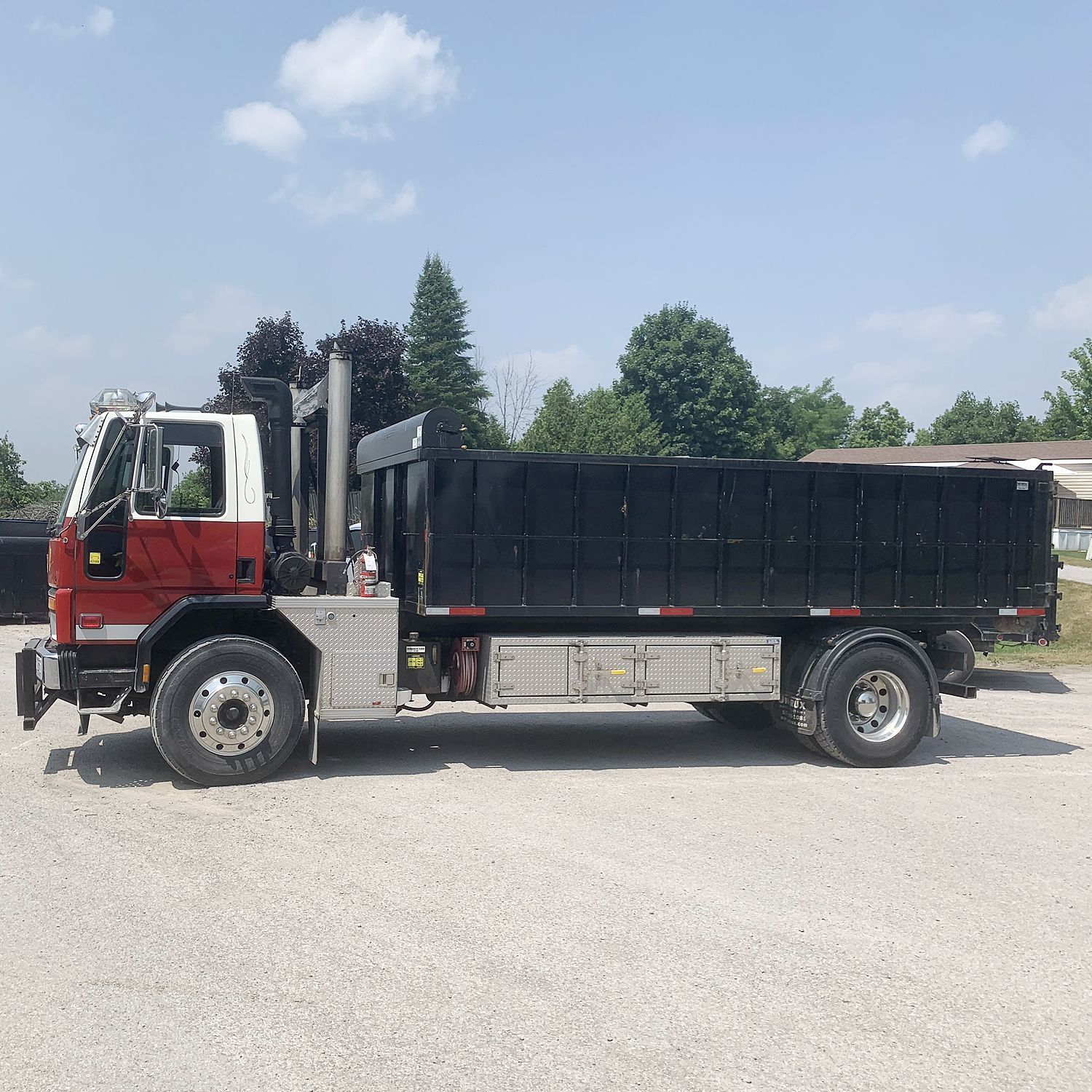 Side view of a red and white cab-over dump truck with a large black dump bed parked on a gravel lot.