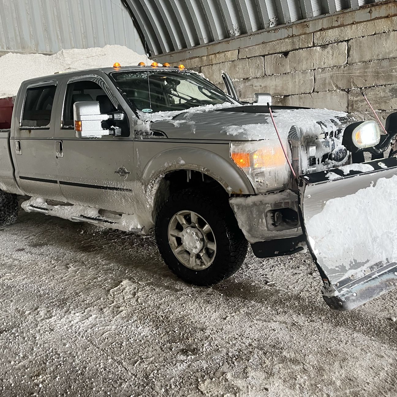 A silver Ford Super Duty pickup truck with a large snowplow attached to the front, parked indoors on a salt-covered floor.